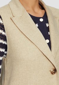 Beige Wolle Weste mit schmalem Revers, ausgestattet mit einem einzelnen goldenen Knopf. Ein dunkelblauer Pullover mit weißen Streifen ist teilweise darunter sichtbar.
