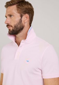 Lichtroze polo shirt met een verhoogde kraag, twee knopen en een klein geborduurd logo met een blauw en roze ontwerp op de linkerborst.