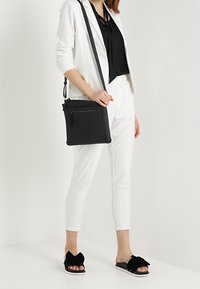 Borsa a tracolla nera con tasca con zip, forma rettangolare, materiale testurizzato e dettagli di cucitura a contrasto, indossata con un completo bianco a righe.