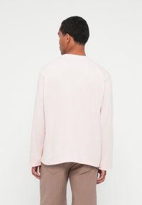 Camicia a maniche lunghe rosa chiaro con scollo rotondo, vestibilità rilassata e texture morbida, abbinata a pantaloni beige a costine.