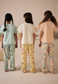 Kolme lasta seisoo rivissä, päällään vaaleansävyiset T-paidat, joissa on röyhelöhihoja, ja kukkakuosiset pyjamahousut eri väreissä.