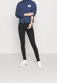 Granatowa kurtka przeciwwiatrowa z zamkiem i łatką z logo, czarne dopasowane legginsy oraz białe sneakersy z jasnymi akcentami. Gładkie, lekkie tkaniny.