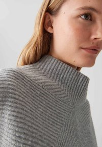 Nahaufnahme einer Person, die einen hellgrauen, gerippten Strickpullover mit hohem, umgeschlagenem Kragen trägt.