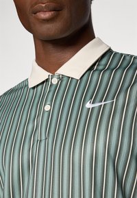 Primer plano de un hombre de piel oscura que lleva una camiseta polo de Nike de rayas verticales verdes y blancas, con cuello y botones blancos.