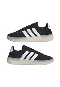 Scarpe sportive nere con design a tre strisce bianche, materiali in suede e sintetico, punta arrotondata e suola in gomma semi-trasparente.