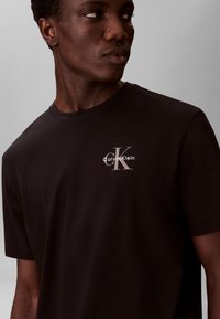 Jeune homme portant un t-shirt noir Calvin Klein regardant sur le côté devant un fond gris uni.