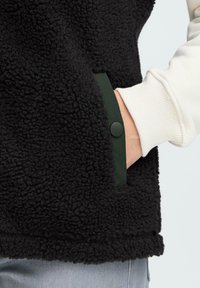 Hand in een zwarte, gestructureerde fleece jaszak met een donkere drukknoop, met een lichtcrème geribde sweatshirtmouw en lichtblauwe spijkerbroek.