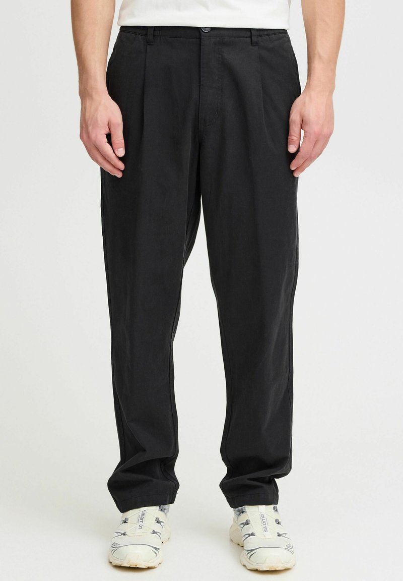 Pantalones amplios de color negro, hechos de una tela ligera, con pliegues frontales, cierre de botón y bolsillos laterales. Combinados con zapatillas blancas.