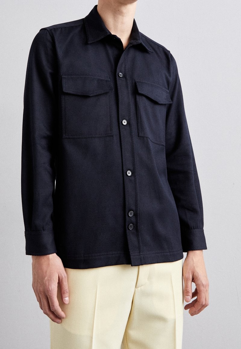 Dark blue shirt with long sleeves, two chest pockets, button closure, and a collar. Features a textured fabric and straight hem.