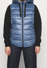 Padded vest w kolorze jasnoniebieskim, z zapięciem na zamek, kapturem ze sznurkiem ściągającym i czarnymi akcentami. Gładka tekstura z poziomym pikowaniem.