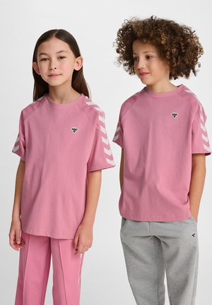 Dwoje dzieci nosi t-shirty z okrągłym dekoltem w miękkim różowym kolorze z bawełny. Jeden z nich ma białe paski w zygzak na rękawach; oba mają małe logo na klatce piersiowej.