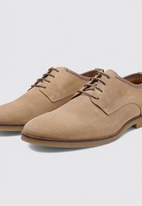 Zapatos de vestir de ante beige con una textura lisa, puntera redondeada y cordones marrones oscuros. Presentan un diseño clásico con acentos mínimos.