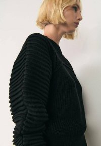 Maglione nero lavorato a maglia con maniche testurizzate e a coste. La forma è aderente nel corpo e ampia nelle maniche, mettendo in risalto dettagli di design unici.