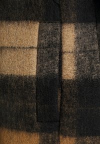 Tessuto in lana a quadri marrone e nero con morbide toppe testurizzate in diverse sfumature, bordate da linee cucite scure. Striscia verticale prominente.