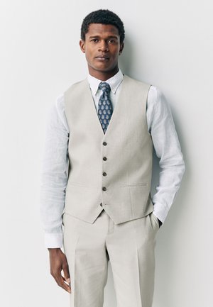 Man in beige suit vest and pants with white shirt and patterned blue tie standing against white wall, hands in pockets.