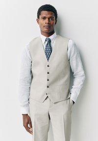Gilet de costume gris clair avec cinq boutons, porté sur une chemise blanche à boutons et une cravate bleue à motifs. Tissu lisse, coupe ajustée, design minimaliste.