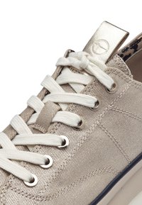 Sneaker beige in tela con lacci bianchi, dettagli dorati metallici e logo circolare sul tallone, caratterizzata da un tessuto testurizzato e occhielli.