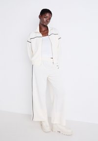 Ensemble de survêtement blanc avec des bordures noires ; veste zippée à col montant et pantalon large. Le modèle porte un haut blanc ajusté et des chaussures blanches épaisses.