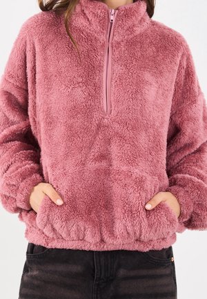 Femme portant un pull duveteux rose à demi-fermeture éclair, les mains dans les poches avant et un pantalon noir visible de la taille vers le bas.