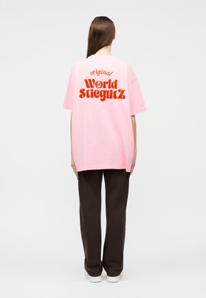 Oseba z dolgimi lasmi, oblečena v dolgo roza majico z rdečim napisom »original World of Stieglitz« na hrbtni strani, temne hlače in bele čevlje.