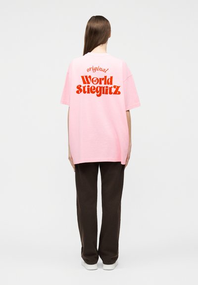 Άτομο με μακριά μαλλιά που φοράει ροζ φαρδιά μπλούζα με κόκκινο κείμενο "original World of Stieglitz" στην πλάτη, σκούρο παντελόνι και λευκά παπούτσια.