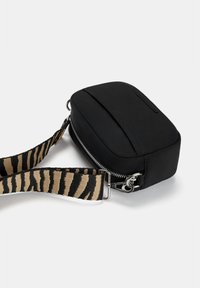 Sort nylontaske med rektangulær form, udstyret med lynlås, og en justerbar rem i stribet beige og sort. Glat, blød tekstur.