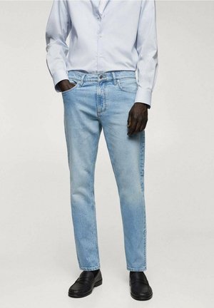 Jambes portant un jean bleu clair coupe droite, des mocassins noirs et une chemise blanche à manches longues boutonnée, une main dans la poche.