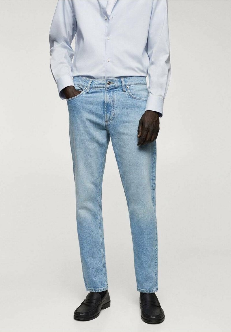 Jambes portant un jean bleu clair coupe droite, des mocassins noirs et une chemise blanche à manches longues boutonnée, une main dans la poche.