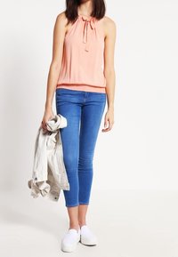 Top de tirantes melocotón con cuello anudado, combinado con jeans ajustados azules y zapatos blancos sin cordones, sosteniendo una chaqueta gris claro.