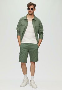 Groene katoenen shirtjacket, witte tanktop, groene cargoshorts, witte sneakers en een zonnebril. Casual fit met knoopzakken op de borst.