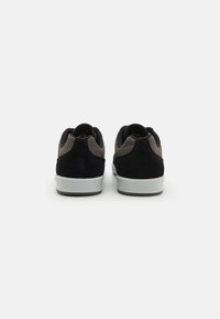Baskets en suede noir et gris avec des accents en mesh, au design arrondi, et semelles en caoutchouc blanches, montrant le talon et la vue arrière.