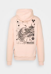 Hoodie rosa chiaro con un'illustrazione grafica nera di una tigre e di una pagoda, circondata da accenti geometrici e testo giapponese.