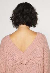 Różowy sweter na drutach z dekoltem w serek, ozdobiony fakturowanym ściegiem i miękkim, przytulnym wyglądem. Tył odsłania skórę i naturalne włosy.