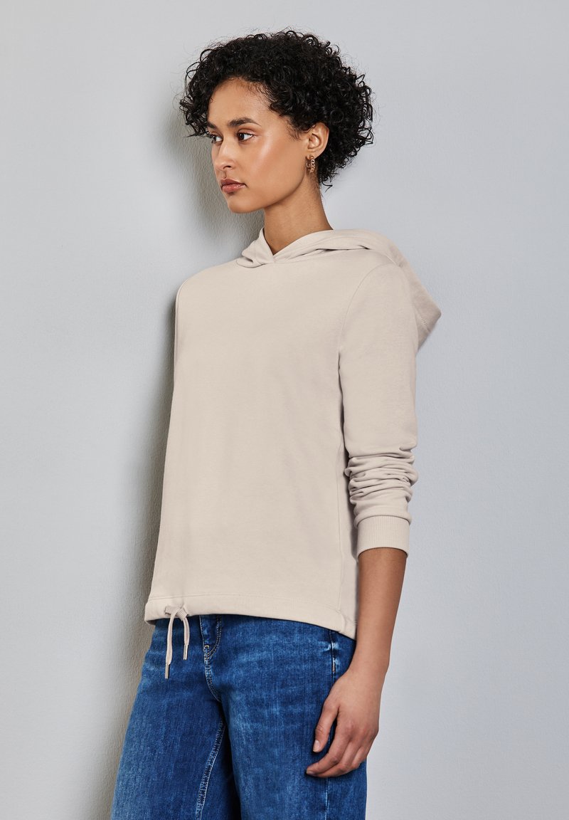 Haut à capuche beige avec un cordon de serrage à l'ourlet, des manches longues et une texture douce. Associé à un jean bleu, offrant une coupe décontractée.