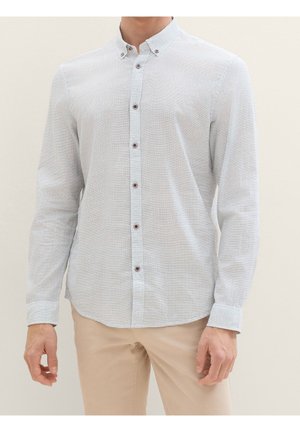 Homme portant une chemise à manches longues bleu clair avec des rayures horizontales subtiles et un pantalon beige, sur un fond neutre.