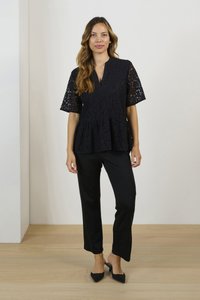 Top peplum in pizzo nero con scollo a V e maniche corte, abbinato a pantaloni neri a gamba dritta e decolleté neri a punta su un pavimento in legno.