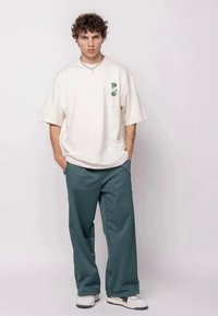 Jovem com cabelo encaracolado a usar uma t-shirt branca oversized com estampado gráfico, calças largas azuis, sapatilhas brancas e um colar de corrente prateado.