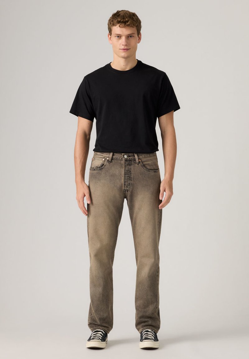 Joven de pie de frente, vestido con una camiseta negra, vaqueros marrones desgastados y zapatillas negras y blancas, sobre un fondo liso.