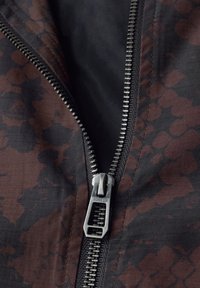 Nahaufnahme einer teilweise geöffneten schwarzen und dunkelroten Jacke mit Muster, die einen Metallreißverschluss mit der Aufschrift "STREET ONE" hat.