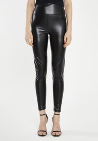 Leggings neri a vita alta realizzati in un materiale lucido e elastico; design aderente con texture liscia; abbinati a scarpe con tacco a caviglia nere.