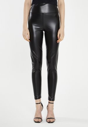 Leggings neri a vita alta realizzati in un materiale lucido e elastico; design aderente con texture liscia; abbinati a scarpe con tacco a caviglia nere.