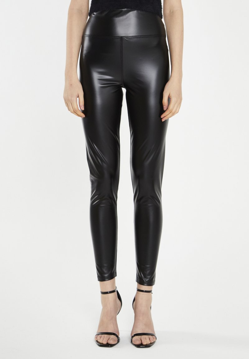 Leggings neri a vita alta realizzati in un materiale lucido e elastico; design aderente con texture liscia; abbinati a scarpe con tacco a caviglia nere.