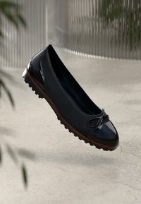 Black ballet flats with a glossy toe cap, a textured upper, decorative bow, and a red-accented sole featuring traction grooves.