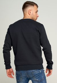 Dunkelgrauer Sweatshirt mit Rundhalsausschnitt, gerippten Bündchen und Saum. Hat einen kleinen Akzent am linken Ärmel. Getragen mit ausgefransten blauen Jeans.