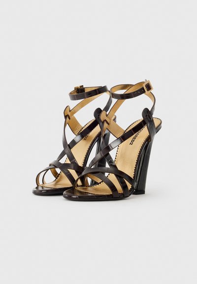 Black strappy high-heeled sandals with a glossy finish, gold accent straps, and a transparent block heel. Open toe design with a textured insole.