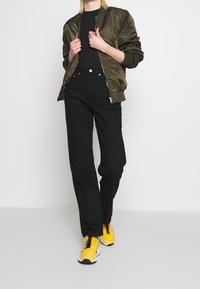 Giacca bomber verde oliva, top nero aderente, pantaloni neri a vita alta e sneakers gialle con dettagli neri. Design semplice con tessuto testurizzato.