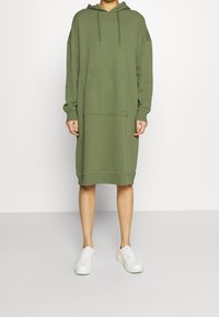 Abito felpa verde con cappuccio realizzato in materiale morbido; presenta una tasca frontale e polsini a coste; abbinato a sneaker bianche.