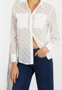 White sheer button-up shirt featuring dual chest pockets, intricate patterned fabric, and a classic collar. Styled with dark blue jeans.