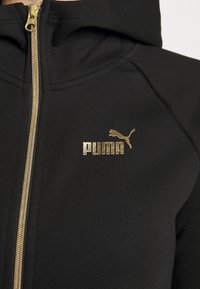 Sweat à capuche noir zippé avec une fermeture éclair dorée et un logo Puma doré sur la partie gauche de la poitrine.