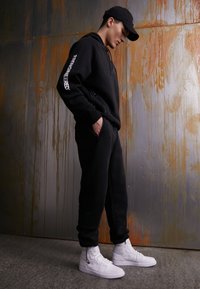 Svart hoodie med vitt logotyp på ärmen, matchande joggers och vita högtoppsneakers. Slät tyg, avslappnad passform, står mot en texturerad bakgrund.